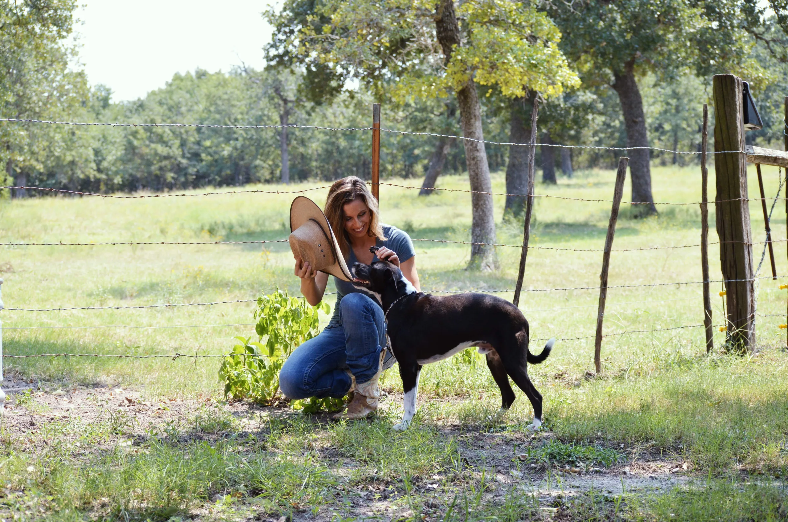 Woman kneeling with fedora and cattle dog in a grassy field with foliage and trees around.