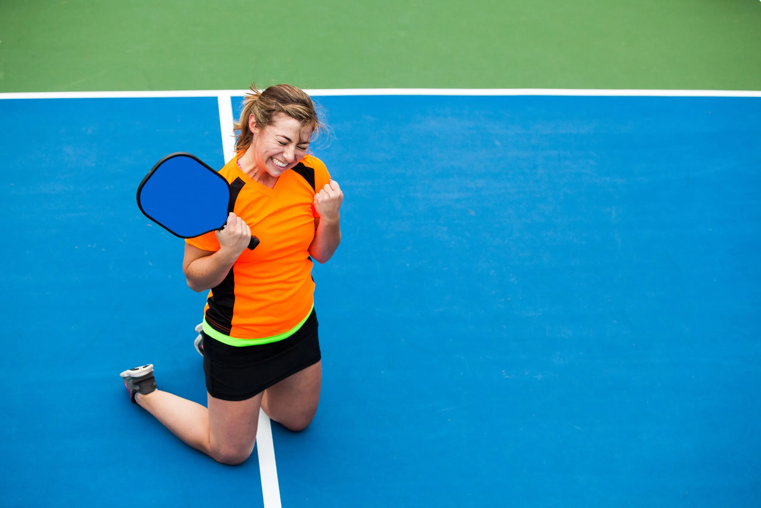 Girl wincing in victorious pose on her knees on a pickleball court after winning pickleball tournament. she has her racket in one hand and her other is a fist held closely to her. She is wearing an orange and black athletic tee and tennis skirt and sneakers.
