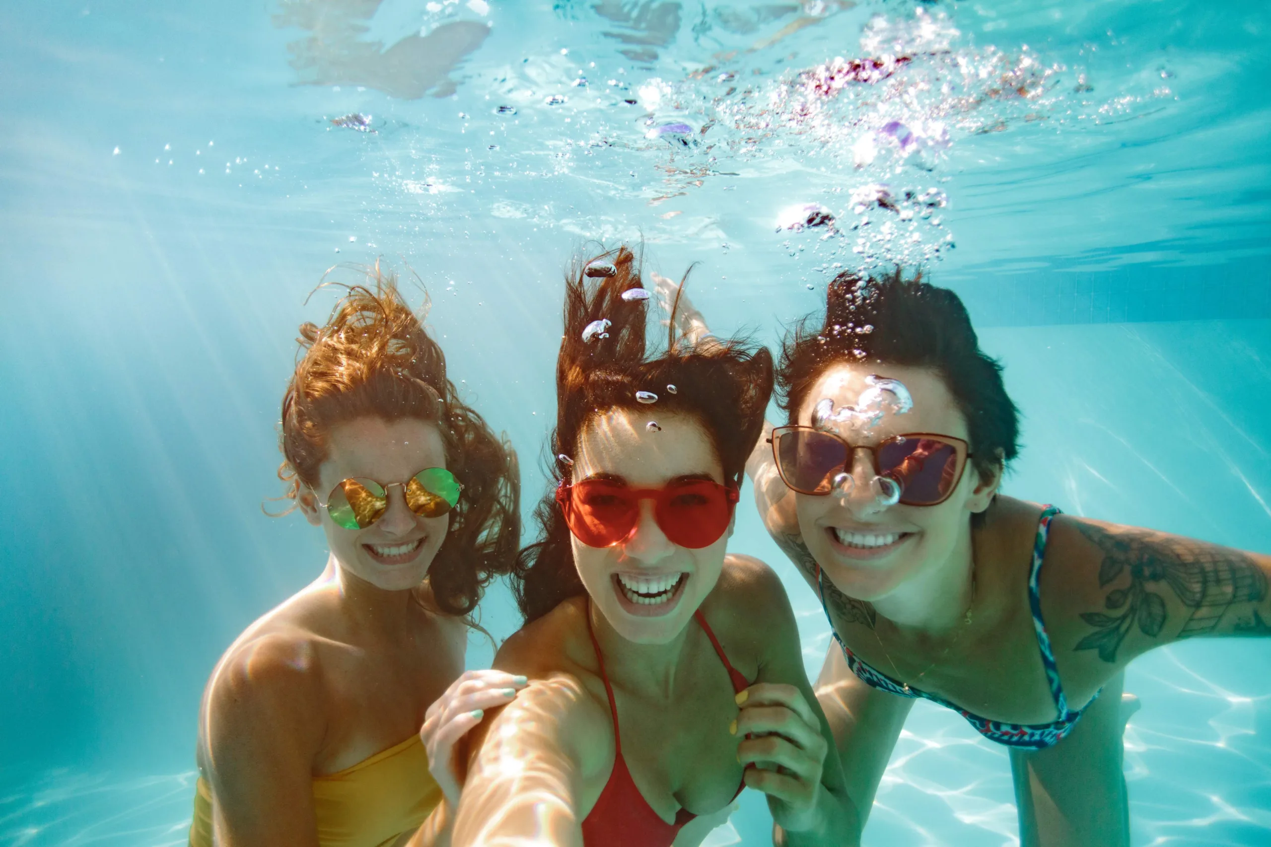 Three young women who are close friends take a selfie underwater with sunglasses on and bubbles coming out of their mouths from smiling and laughing. The woman on the left has gold sunglasses on and wearing a mustard yellow swimsuit. She is holding the middle woman's arm who is taking the picture with her right arm and has red sunglasses and a red bikini on. The third woman is on the far right and in a blue and white abstract pattern bikini with brown sunglasses on and a left arm tattoo of a plant with flowers and a bird cage.
